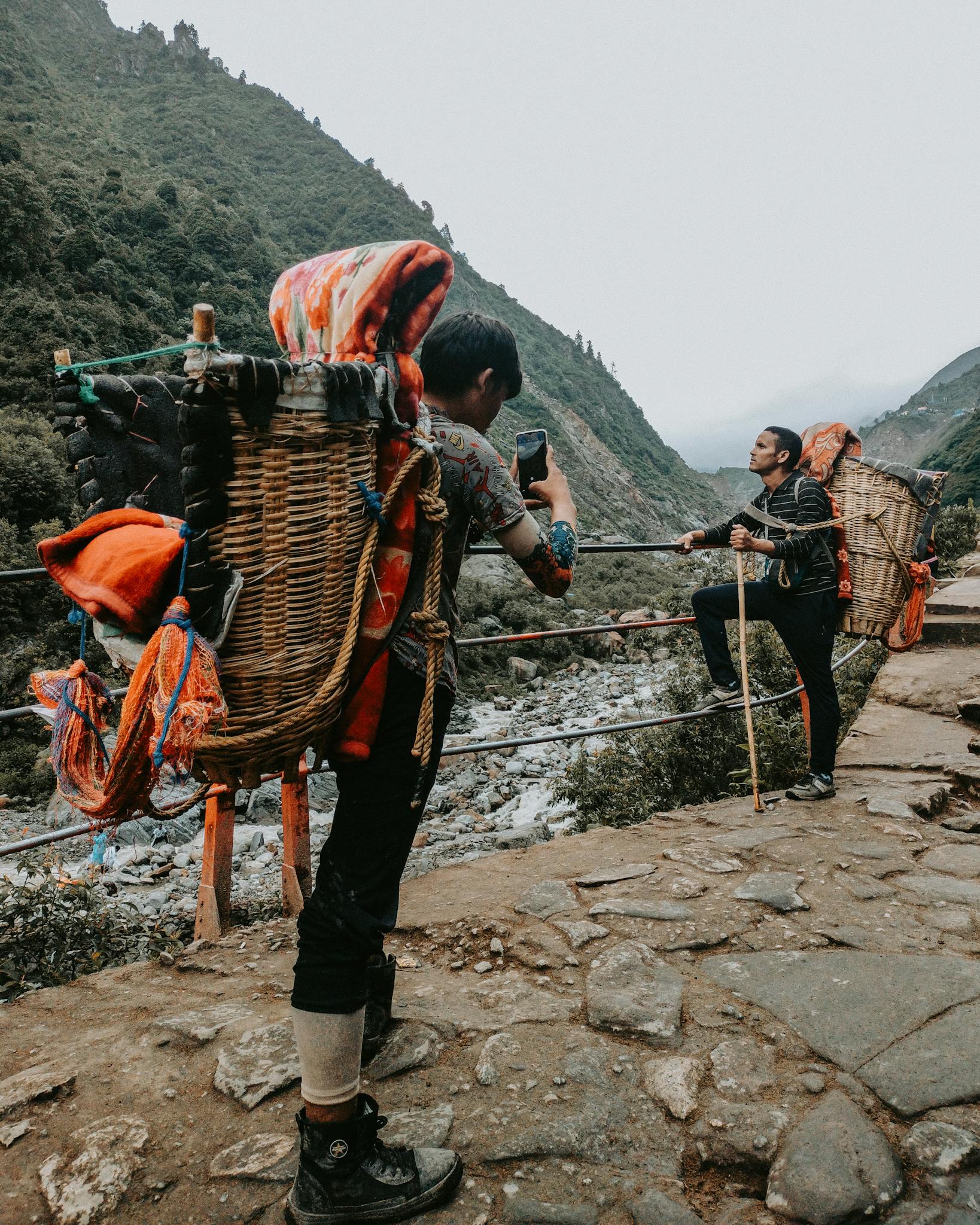 Two men with traditional backpacks trek a scenic mountain path, capturing a moment with a smartphone.